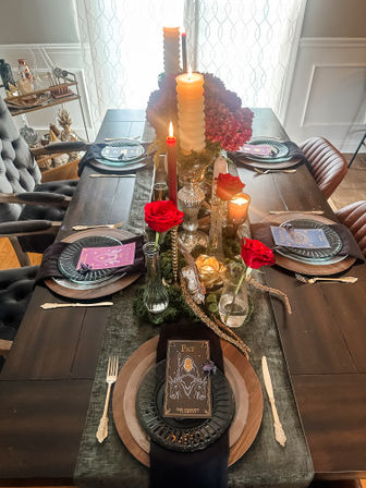 Candlelit dark-wood dining table tablescape with layered plates and tarot-style place cards, red roses in glass vases, tall ribbed candles, and a lush hydrangea-and-moss centerpiece in a cozy dining room.