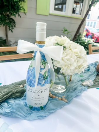 Outdoor garden table centerpiece: wine bottle with blue hydrangea label tied with a white bow beside a white hydrangea bouquet on a pale blue table runner.