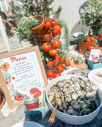 Sunlit outdoor table with vine-ripened cherry tomatoes in a wire basket, potted herbs, a framed menu, jar of tomato jam and a bowl of steamed clams — a bright summer seafood spread.