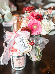 Festive table centerpiece: gift-wrapped bottle of pink prosecco tied with pink tulle and ribbon beside a vase of pink gerbera, red and white roses, eucalyptus, and small mirrored disco ball ornaments.