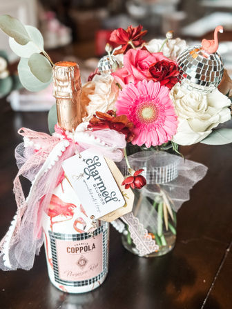 Festive table centerpiece: gift-wrapped bottle of pink prosecco tied with pink tulle and ribbon beside a vase of pink gerbera, red and white roses, eucalyptus, and small mirrored disco ball ornaments.