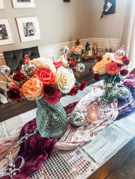 Dining room table centerpiece with two textured glass vases of mixed roses, gerbera and carnations in coral, pink and white, accented by sparkling mini disco-ball ornaments, a lit votive and layered mauve and burgundy fabric runners on a mirrored table runner.