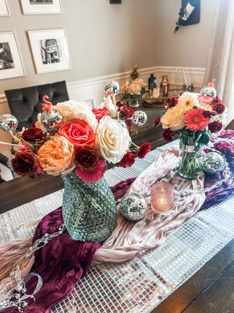 Dining room table centerpiece with two textured glass vases of mixed roses, gerbera and carnations in coral, pink and white, accented by sparkling mini disco-ball ornaments, a lit votive and layered mauve and burgundy fabric runners on a mirrored table runner.