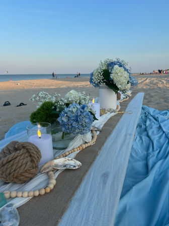 Coastal beach table setting on sandy shore with blue and white hydrangeas, glowing candles, shells and nautical rope overlooking the calm ocean at sunset