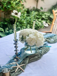Outdoor garden patio table centerpiece with white hydrangeas and anemone blooms in glass vases, silver candlestick, blue-green fabric runner and decorative starfish — coastal-inspired table decor.