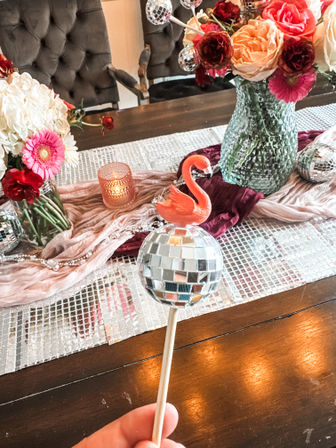 Playful disco-ball wand topped with a pink flamingo held above a dining table centerpiece of mixed roses and gerbera in glass vases, mirrored runner and candlelight