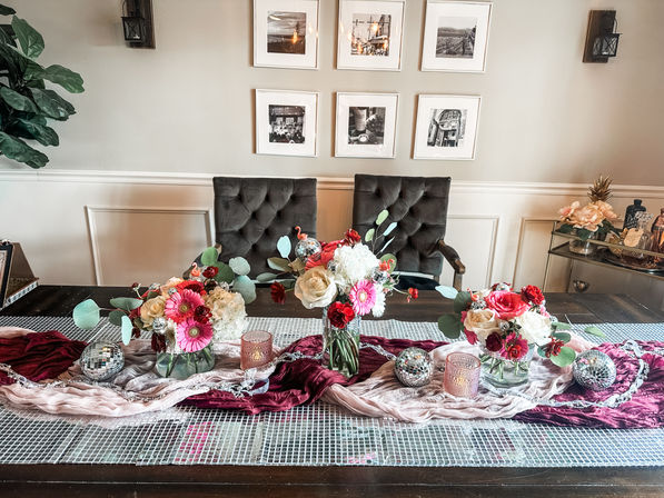 Dining room table centerpiece with pink, red and white floral arrangements in glass vases, burgundy and blush fabric runner, small disco-ball accents and candles, tufted dining chairs and framed photos on a beige wall.