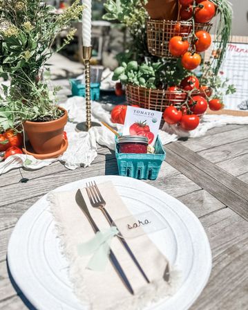 Al fresco wooden table with a white charger, frayed linen napkin and tied cutlery with a small place card; centerpiece of potted herbs, hanging cherry tomatoes in wire baskets, a brass candle, and a teal berry box holding a jar and tomato seed packet — bright summer garden table setting.
