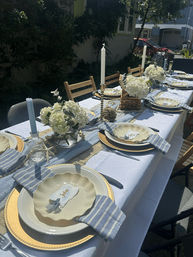 Backyard summer table set for a garden party — blue-and-white striped napkins, scalloped cream plates on gold chargers, hydrangea centerpieces, glass candlesticks with white tapers and a rustic table runner.