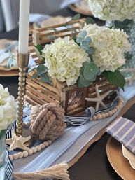 Nautical coastal centerpiece with white hydrangeas and eucalyptus in a rustic wooden crate, knotted rope, starfish, striped navy ribbon, wooden bead garland and gold candlestick on a blue table runner.