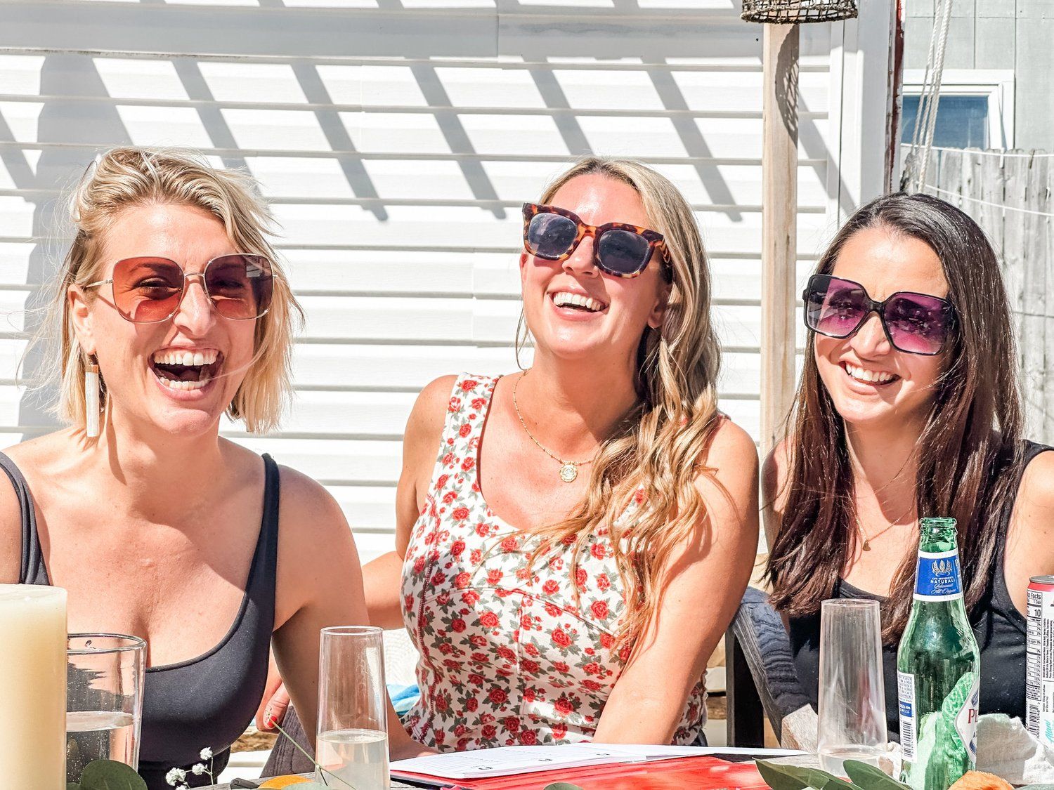Three smiling women in sunglasses laughing at a sunny outdoor patio brunch with drinks on the table, summer casual gathering