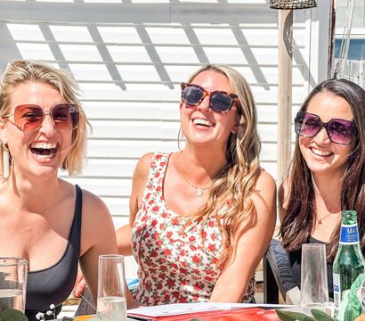Three smiling women in sunglasses laughing at a sunny outdoor patio brunch with drinks on the table, summer casual gathering