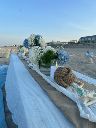 Coastal-chic beach table on sandy shore with burlap runner, white and blue hydrangea centerpieces, baby's breath, a glass candle, nautical rope knot and starfish, oceanfront buildings under a clear sky.