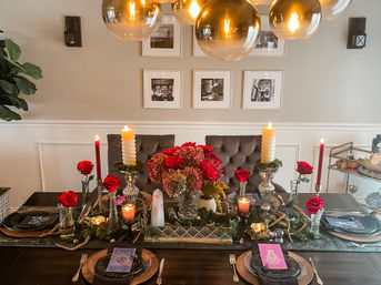 Elegant indoor dining room table set for dinner with a lush centerpiece of red roses and hydrangeas, tall twisted and taper candles in crystal holders, black plates on copper chargers, tufted velvet chairs, gold globe pendant lights overhead, and framed black-and-white photos on a beige wall.