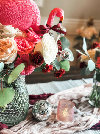 Indoor dining table floral centerpiece with a pink plastic flamingo, mixed roses and gerbera, eucalyptus in a textured glass vase, small disco ball ornaments and a lit pink votive on a velvet runner.