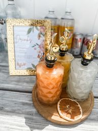 Sunlit outdoor brunch setup: three patterned glass pour bottles with gold spouts holding orange, yellow, and white cocktail mixers on a round wooden tray with a dried citrus slice, framed floral drinks sign and beverage bottles in the background.