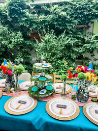 Outdoor backyard garden party table with turquoise tablecloth, gold-rimmed chargers, layered blush plates with tassel place cards, moss-covered tiered dessert stand with blue cookies, and bright floral centerpieces in front of a leafy trellis.