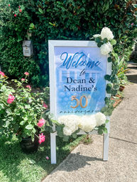 Sunny front-yard 50th anniversary welcome sign on a white easel with blue watercolor print, draped in white hydrangeas, eucalyptus and pink roses by a sidewalk
