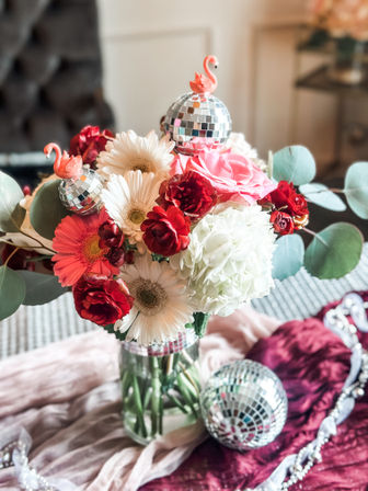 Indoor floral centerpiece in a glass vase with pink roses, white gerbera daisies, red ranunculus, white hydrangea and eucalyptus, accented by sparkly disco ball ornaments and tiny pink flamingo toppers on a velvet table runner.