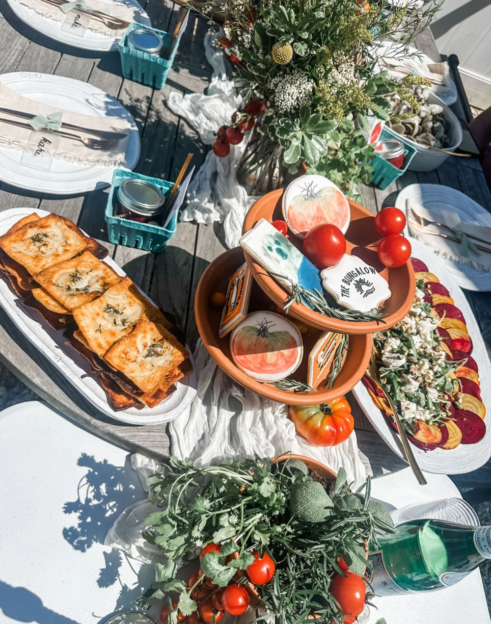 Outdoor summer brunch spread on a wooden patio table with fresh herbs and cherry tomatoes, terracotta bowls of tomatoes and decorated cookies, golden puff pastries, a beet and goat cheese salad, jars and sparkling water, and a wildflower centerpiece