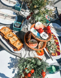 Outdoor summer brunch spread on a wooden patio table with fresh herbs and cherry tomatoes, terracotta bowls of tomatoes and decorated cookies, golden puff pastries, a beet and goat cheese salad, jars and sparkling water, and a wildflower centerpiece
