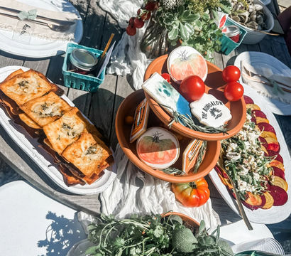 Outdoor summer brunch spread on a wooden patio table with fresh herbs and cherry tomatoes, terracotta bowls of tomatoes and decorated cookies, golden puff pastries, a beet and goat cheese salad, jars and sparkling water, and a wildflower centerpiece