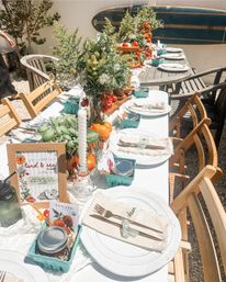 Sunny outdoor patio tablescape for a summer brunch — white-linen table with wildflower centerpieces, heirloom tomatoes, potted basil, candles, mason jars and surfboard backdrop.