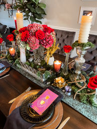Lush indoor dining table centerpiece with red roses and pink hydrangeas, tall cream candles, moss runner, gold votives, black plates and a bright pink tarot-style place card.