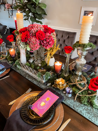 Lush indoor dining table centerpiece with red roses and pink hydrangeas, tall cream candles, moss runner, gold votives, black plates and a bright pink tarot-style place card.