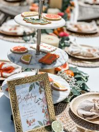 Sunlit citrus-themed birthday brunch table with marble tiered stand of decorated orange and lemon cookies, gold framed sign, woven chargers, and a lime garnish.