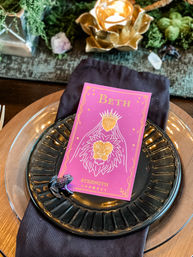 Dining place setting with a pink tarot-style place card reading "Strength" and a gold lion illustration, placed on a black decorative plate over a dark napkin and clear charger, with a small frog figurine and candlelit crystal decor on a wooden tablescape.