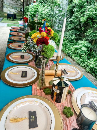 Backyard garden party table setting with teal tablecloth, gold chargers, blush plates with numbered place cards, pink gauzy runner, bright mixed-flower centerpieces, brass candlestick with white taper, and a decorative 'secrets' jar.
