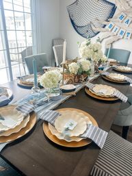 Sunlit coastal-themed dining table set for a seaside birthday with white hydrangea centerpieces, blue taper candles, striped napkins, scalloped plates on gold chargers, nautical net wall decor and deck view.