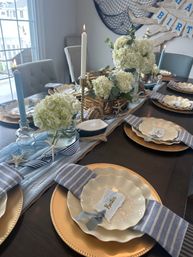 Beachy coastal dining table set for a celebration with white hydrangea centerpieces, starfish and rope accents, gold charger plates, scalloped white dishes, striped blue napkins and lit taper candles in a bright dining room