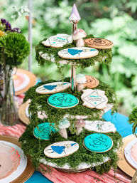 Three-tier moss-covered dessert stand outdoors styled as a whimsical garden party centerpiece with decorated sugar cookies (teal rune designs, illustrated girl in blue dress), tiny mushroom toppers and pastel accents.