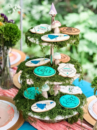 Three-tier moss-covered dessert stand outdoors styled as a whimsical garden party centerpiece with decorated sugar cookies (teal rune designs, illustrated girl in blue dress), tiny mushroom toppers and pastel accents.