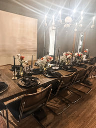 Spooky-chic indoor dining table set for a Halloween dinner — long wooden table with gold-rimmed black chargers, black napkins, tall black candles, skull accents and peach-and-white floral centerpieces under a modern chandelier.