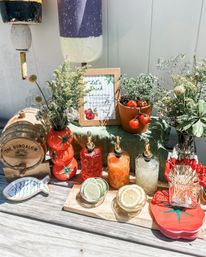 Outdoor patio summer cocktail station with a small wooden barrel, three decorative glass pour bottles, bowls of lime and lemon slices, potted cherry tomatoes and fresh herb and floral arrangements