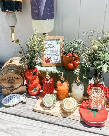 Outdoor patio summer cocktail station with a small wooden barrel, three decorative glass pour bottles, bowls of lime and lemon slices, potted cherry tomatoes and fresh herb and floral arrangements