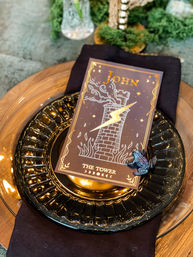 Mystical tarot-themed dinner place setting featuring a 'The Tower' card with a lightning-struck tower, glossy black glass charger over a clear gold charger, black linen napkin, and a small decorative frog pin on a wooden table.