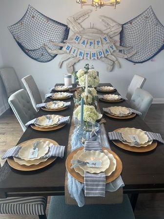 Nautical-themed birthday table in a coastal-style dining room with crab wall art and nets, long dark wood table set with scalloped plates, striped napkins, gold chargers and hydrangea centerpieces.