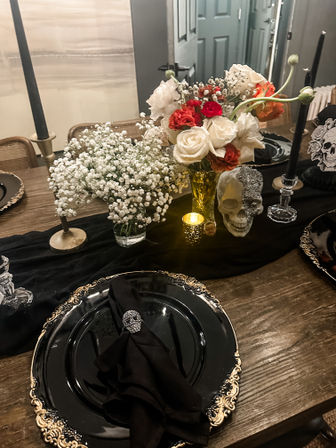 Gothic Halloween tablescape on a rustic wooden dining table: black and gold charger plates with black napkins and skull napkin rings, skull centerpiece with glitter accent, glass vases of white roses, red blooms and baby’s breath, black taper candles and a glowing votive.