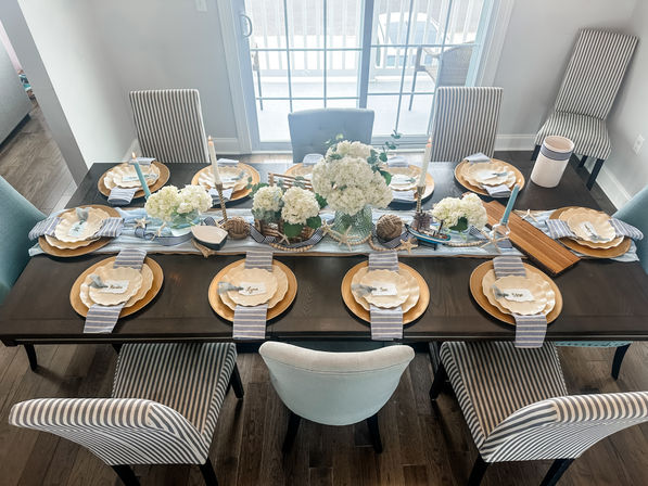 Coastal-inspired dining room table set for eight with gold chargers, scalloped plates, blue-striped napkins, white hydrangea centerpieces, candles and nautical decor by sliding glass doors