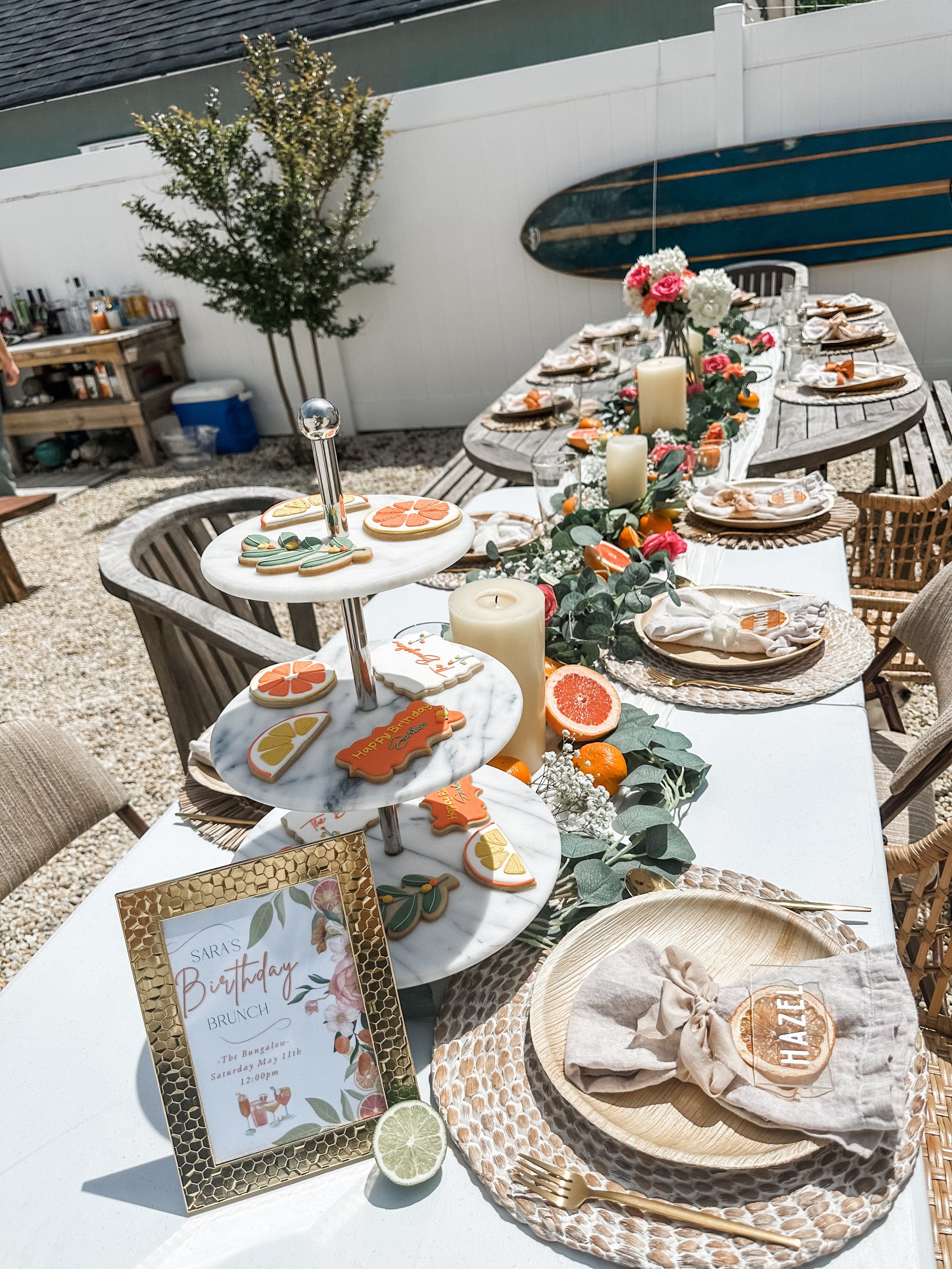 Sunlit backyard patio birthday brunch table with citrus-themed cookies on a marble tiered stand, halved grapefruits and oranges, pillar candles and eucalyptus-rose garland, woven rattan chargers, linen napkins and gold flatware, surfboard propped on a white fence for a coastal vibe.