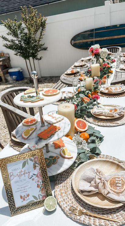 Sunlit backyard patio birthday brunch table with citrus-themed cookies on a marble tiered stand, halved grapefruits and oranges, pillar candles and eucalyptus-rose garland, woven rattan chargers, linen napkins and gold flatware, surfboard propped on a white fence for a coastal vibe.