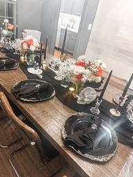 Halloween-themed dining table on a rustic wooden table with black and gold place settings, black taper candles, skull accents and white and orange floral centerpieces.