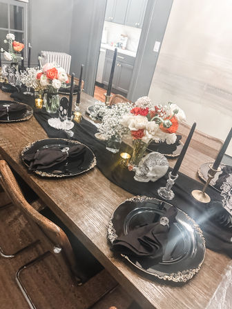 Halloween-themed dining table on a rustic wooden table with black and gold place settings, black taper candles, skull accents and white and orange floral centerpieces.