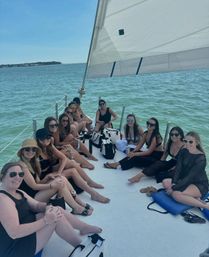 Group of women relaxing on the deck of a white sailboat under a large white sail, turquoise coastal waters and distant shoreline on a sunny summer day.