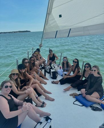 Group of women relaxing on the deck of a white sailboat under a large white sail, turquoise coastal waters and distant shoreline on a sunny summer day.