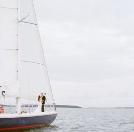 Romantic bride and groom embrace on the bow of a sailboat with a tall white sail, cruising calm coastal waters under an overcast sky.